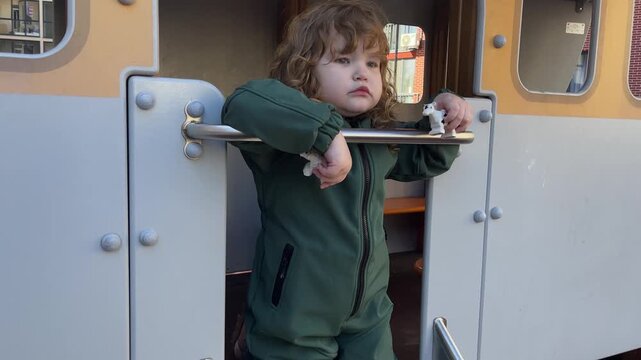 Curly hair child playing on slide at playground. Excited kid preparing for descent on metal chute. Active toddler enjoying outdoor game in public park setting.