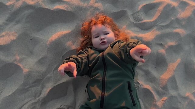 Sad ginger kid lying on sand at beach. Young child with curly hair feeling grumpy during outdoor playtime. Childhood emotions, expression of melancholy and unhappiness in nature.