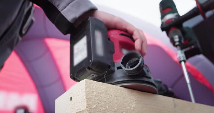 Close-up of a worker's hand carpenter operating a professional red power sander tool on a piece of timber wooden plank. Woodworking process, home renovation, and construction concept.