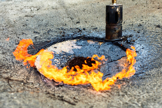 Flames emerge in circular pattern around roof drainage pipe on a damaged rooftop surface. Fire burns intensely around metal drain opening, creating ring of flames on cracked roofing material