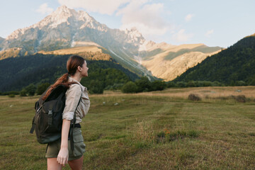 Naklejka premium A woman hiker with a backpack stands in an open meadow, facing distant mountains, capturing a moment of outdoor adventure and solo nature travel. In bright daytime scenery.