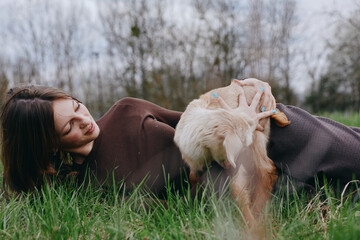 Naklejka premium Woman lying on grass in a cosy sweater while a goat kid nudges her hand, capturing warmth, companionship, and rustic charm in an outdoor setting. Natural light creates a calm, lifestyle mood.
