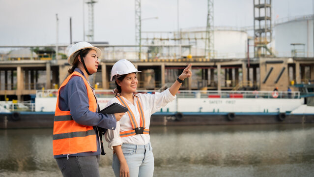 Two engineers stand by water near industrial structures. They wear hard hats and high-visibility vests. One gestures toward a facility while holding a tablet. Their expressions show focus and teamwork