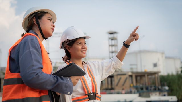 Two engineers in hard hats point at a construction site. They wear high-visibility vests and hold tools. One smiles while the other concentrates on their task. Background shows industrial structures