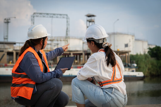 Two engineers in hard hats discuss plans beside a waterway. They point at a tablet while crouching near industrial tanks. The sky shows clouds above distant factory structures
