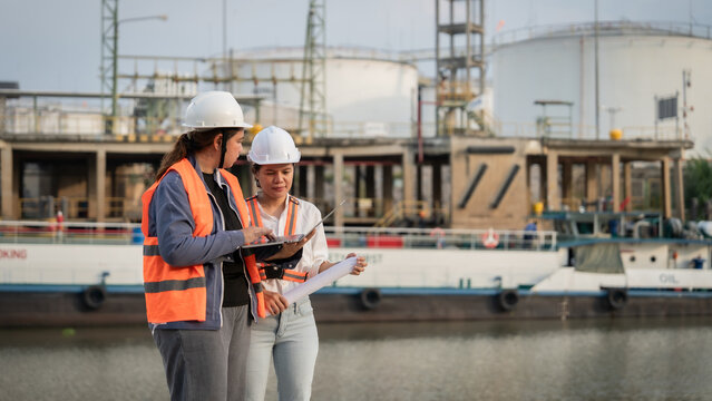 Two engineers stand by water, reviewing plans. White helmets and vests mark their professional gear. Industrial tanks rise behind them, near a docked boat. They focus intently, showing teamwork