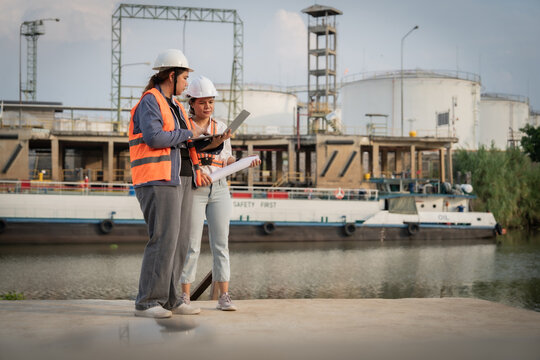 Engineers walk near industrial facility by river. They discuss plans, holding documents together. White helmets and bright vests mark safety gear. Large tanks and structures loom behind them