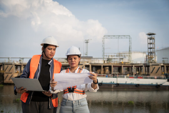 Two engineers stand by a waterfront facility. They wear hard hats and safety vests. One holds blueprints, the other a laptop. They appear focused and collaborative. This image suits industrial