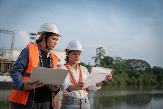 Two engineers review plans near a riverbank. They wear hard hats and safety vests. One holds a laptop, the other holds blueprints. Their focus suggests teamwork and planning