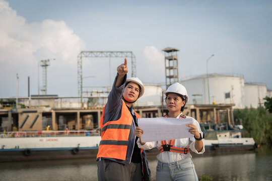 Two engineers inspect an industrial site with focus. They point at blueprints, analyzing structural plans. Calm river flows beside their site. Background towers hint at complex infrastructure