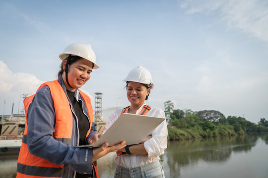 Two engineers stand by a river, discussing plans. One holds a tablet, the other listens attentively. They wear helmets and safety vests for protection. The background shows industrial infrastructure
