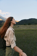 Naklejka premium Young woman with long red hair enjoys a peaceful moment in a vast green meadow surrounded by mountains under a clear blue sky.