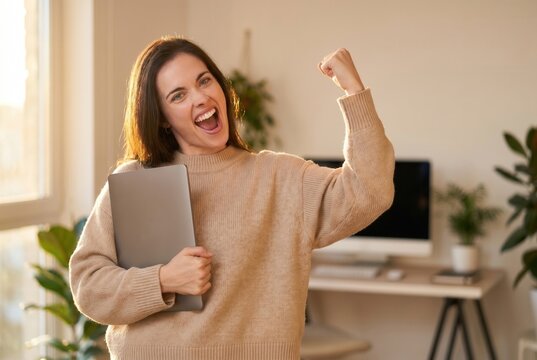 Woman holding a laptop and cheering, feeling success and happiness at her home office