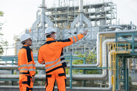 Two engineers industrial workers in orange safety gear discuss tasks at a large chemical or oil refinery plant with complex metal structures and pipelines.