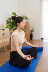 Fototapeten Lotusblume young Asian women practicing yoga meditation lotus pose on blue mat at home living room floor with plants peaceful wellness mindfulness exercise indoor  © snowing12