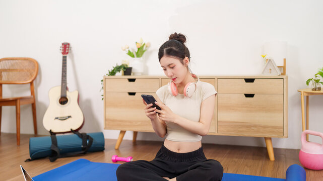 Asian women sitting yoga mat using smartphone mobile phone check fitness app workout home exercise routine living room wellness healthy lifestyle technology