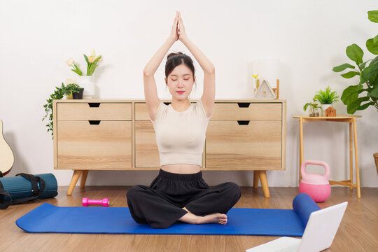 Asian women practicing yoga meditation pose sitting on mat at home living room with laptop online fitness class wellness exercise mindfulness stretching indoor
