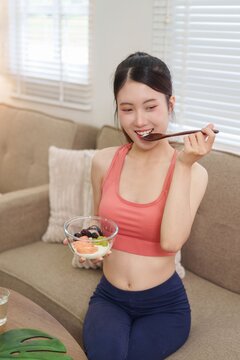 young Asian woman eating healthy yogurt bowl fresh fruit after workout at home living room fit lifestyle wellness nutrition diet breakfast meal fitness