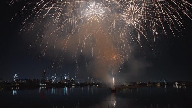 Brilliant fireworks illuminate the night sky over Dubai as golden and white pyrotechnics cascade above the city skyline. Colorful explosions reflect on calm water while smoke drifts across the scene.