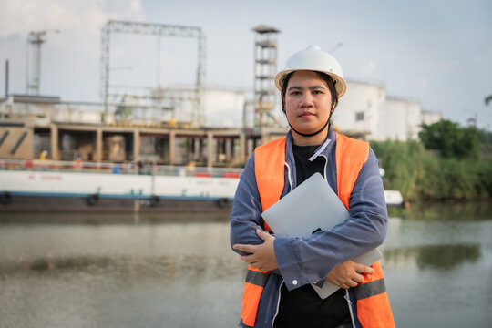 Engineer stands by water near industrial site. Wears hard hat and safety vest. Holds tablet, focused and professional. Background shows construction and storage tanks