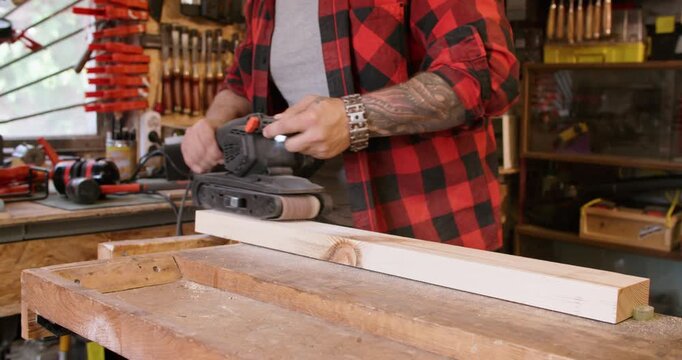 Carpenter occupation. Joinery process. Man smoothing board surface with handheld belt sander in workshop interior with tools on shelves.
