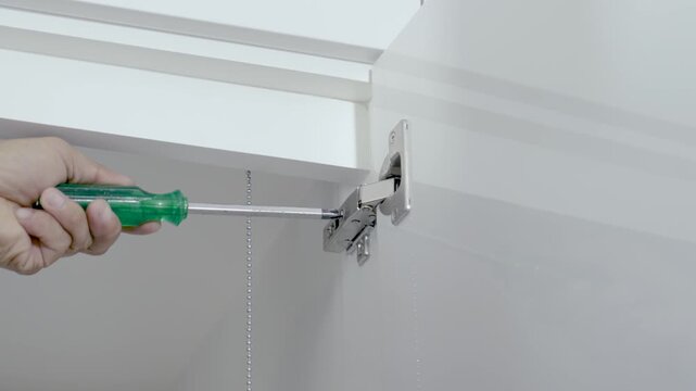 Cabinet assembler uses a screwdriver to adjust the hinge of a white cabinet door.