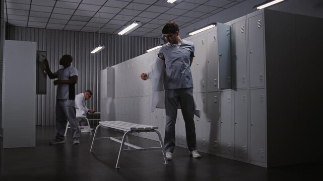 Diverse Groups of Male Doctors in Blue Scrubs and White Coats Undress and Talk by Metal Lockers in Clinic Dressing Room, Finishing Professional Medical Duty. Concept of Healthcare Worker Routine.