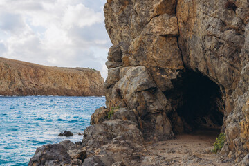 Fototapeta premium Rocky path leading into a natural sea cave on a cliff face, with turquoise Mediterranean Sea and distant headland