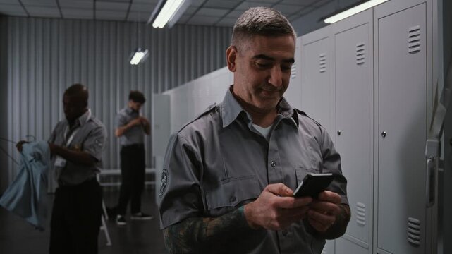 Muscular Male Security Officer With Tattoos at Locker Hangs Up ID, and Uses Smartphone to View Notifications and Social Media After Long Airport Security Shift. Workplace Communication Concept.