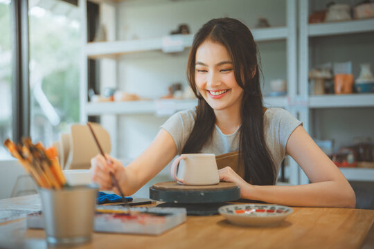 Happy Southeast Asian Gen Z woman smiling while painting a ceramic coffee mug in a pottery studio. A creative female artist enjoying her hobby during a workshop. Concept of mindfulness.