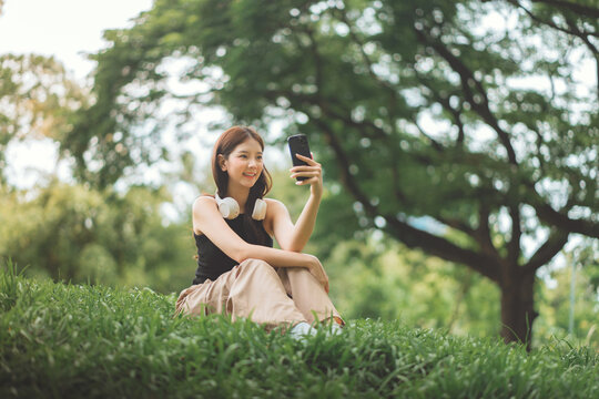 Smiling young Asian woman taking a selfie with a smartphone while sitting on green grass in a lush park. She enjoys a relaxed outdoor lifestyle with headphones around her neck, capturing a happy momen