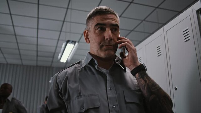 Security Officer With Prominent Arm Tattoos Stands by Grey Metal Locker, Holding Smartphone and Having Serious Conversation After Workday Shift. Workplace Communication Concept in Staff Changing Room.