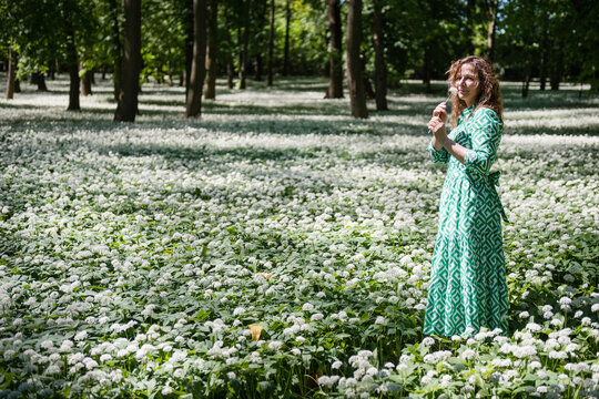 Young smiling woman in a green patterned dress smelling wild garlic flowers (Allium ursinum) while standing in a blooming spring forest park, Dolna Krupa, Slovakia