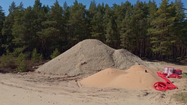 Construction debris scene. Sand and gravel deposits at construction site environment. Scene showing aggregate mounds beside pine forest with construction equipment and sunny weather