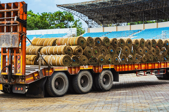 Truck loaded with sod rolls parked at stadium for lawn landscaping and renovation for football field. Large shipment of natural sod rolls on heavy duty truck trailer with stadium bleacher background.