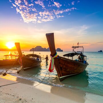 Two wooden boats with ornate prows rest on a sandy shore at sunset. Turquoise waters meet the horizon, silhouetting distant islands. Two people are walking near boats