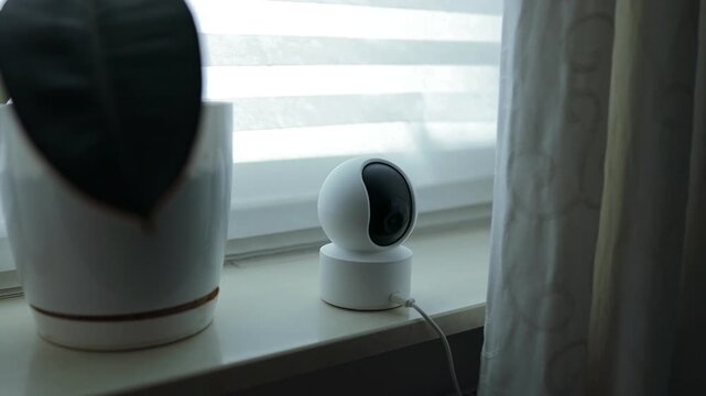 A compact wireless smart home security camera or baby monitor standing on a windowsill in a bright room. This footage captures modern technology used for home surveillance, child monitoring, and