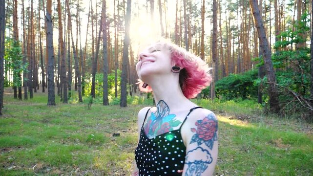 Carefree hippie woman with pink hair having fun at summer forest. Happy smiling punk girl with tattoos fooling around woodland. Young female hipster with ear tunnels enjoying rest at summer nature