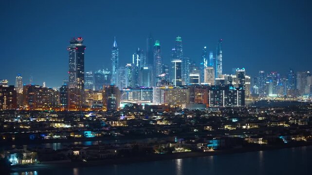 Stunning nighttime aerial view of Dubai Marina skyline with illuminated skyscrapers towering over Palm Jumeirah waterfront villas, city lights reflecting on calm waters under deep blue twilight sky.