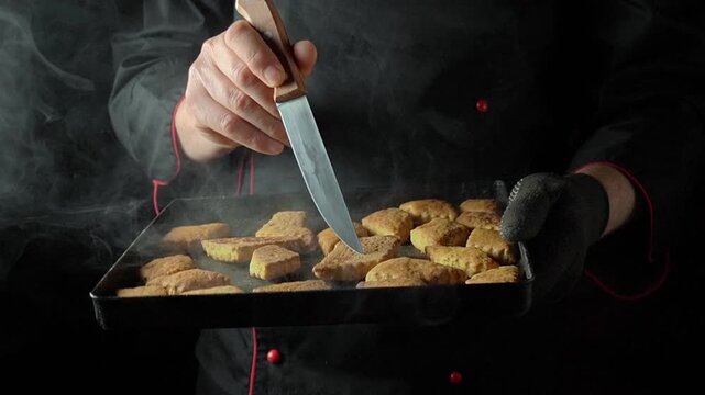Chef in black attire carefully checks freshly baked cookies on a tray, with steam rising in a dark kitchen, showcasing the delicious treats and the meticulous preparation process