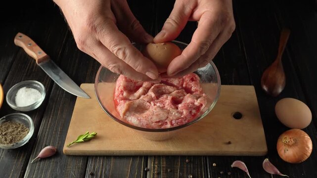 Hands breaking eggs into a bowl of minced meat on a wooden cutting board with kitchen utensils and ingredients arranged around the workspace
