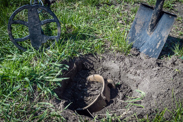 A clay pot from the Scythian period with coins or a hoard in a dug pit. Nearby is a metal detector for searching the ground and a shovel. The area is grassy and sunny, with signs of outdoor activity © Віталій Б.
