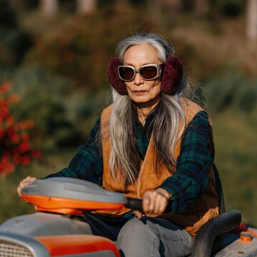 Older woman riding mower through landscaped yard wearing earmuffs