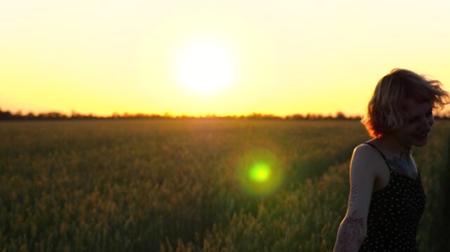 Happy female hipster with tattoos having fun during goes through green barley field at beautiful sunset. Cheerful punk girl in short black dress fooling around while walks among wheat meadow. Slow mo