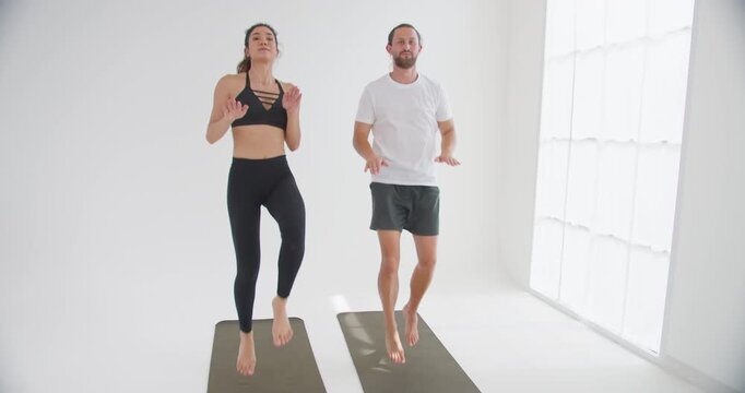 A fit couple performs a synchronized high-knee cardio exercise on yoga mats in a bright white studio, focusing on core strength and physical wellbeing together.