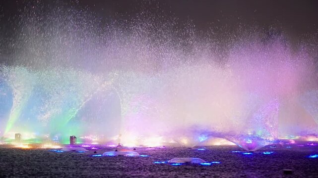 Spectacular nighttime fountain show in Dubai featuring multicolored LED lights illuminating water jets with dramatic fire bursts. Palm trees silhouetted against vibrant purple blue and orange spray.