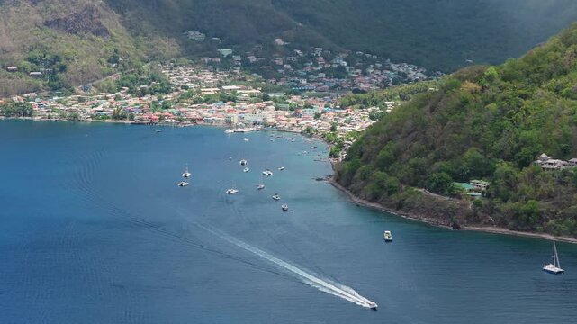 Aerial footage of Soufriere Bay in Saint Lucia featuring colorful Caribbean town nestled between lush green Piton mountains with sailboats and speedboat crossing turquoise waters.