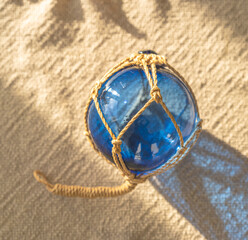 Blue glass fishing float wrapped in rope resting on sand with warm sunlight and coastal shadow patterns. © Trygve