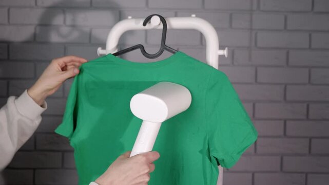 Woman steaming green t-shirt with garment steamer. Close-up of female hands steaming a crumpled green cotton t-shirt with a portable appliance, smoothing out wrinkles for a neat look