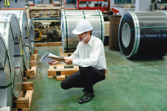 Caucasian man intently reviews technical documentation amidst gray metal sheet factory environment, wearing dark suit and focused expression, examining blueprints with detailed machinery.